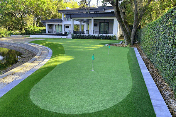 Luxury backyard custom synthetic putting green with multiple holes and flags, professionally installed by JR Landscaping Service near a modern home and pond