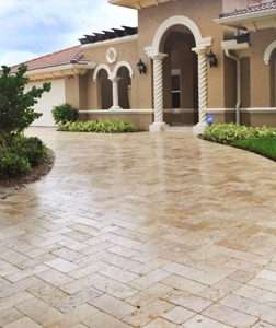 A sweeping driveway featuring light tan travertine pavers laid in a herringbone pattern, leading up to the arched entrance of a Mediterranean-style home in Clearwater, Florida.