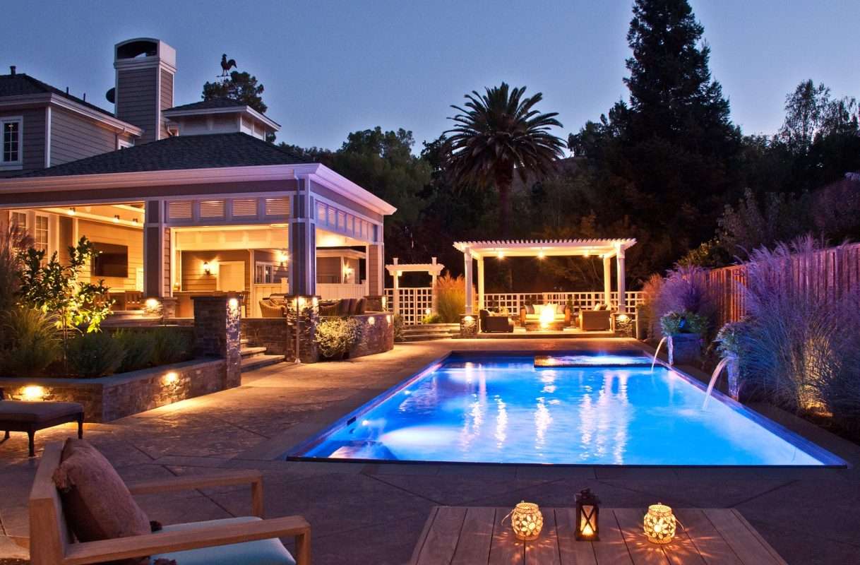 Night view of a St. Petersburg backyard featuring a lit swimming pool, pergola, and landscape lighting accentuating plants and seating areas.