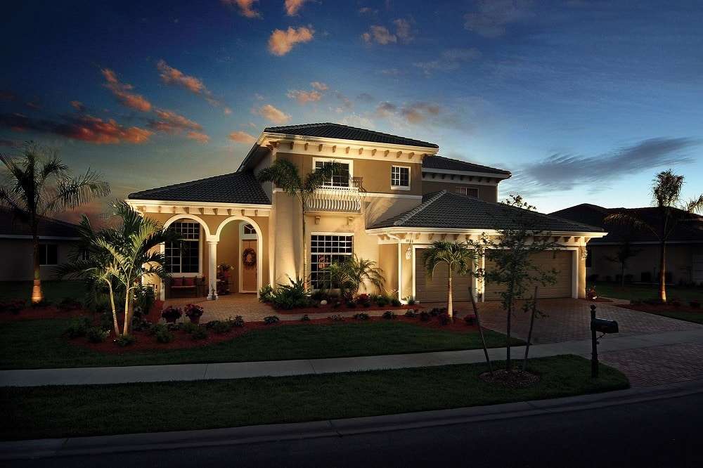 A luxury residence typical of Davis Island’s prestigious streets shown at twilight. Professional-grade brass up-lighting dramatically highlights the textured stucco facade, architectural columns, and mature towering royal palm trees emblematic of the neighborhood. Subtle path lights clearly define manicured walkways and garden beds, while wash lights enhance the lush, dense tropical foliage that lines the property boundary. The lighting design is cohesive, highlighting the property’s value while providing complete visibility.