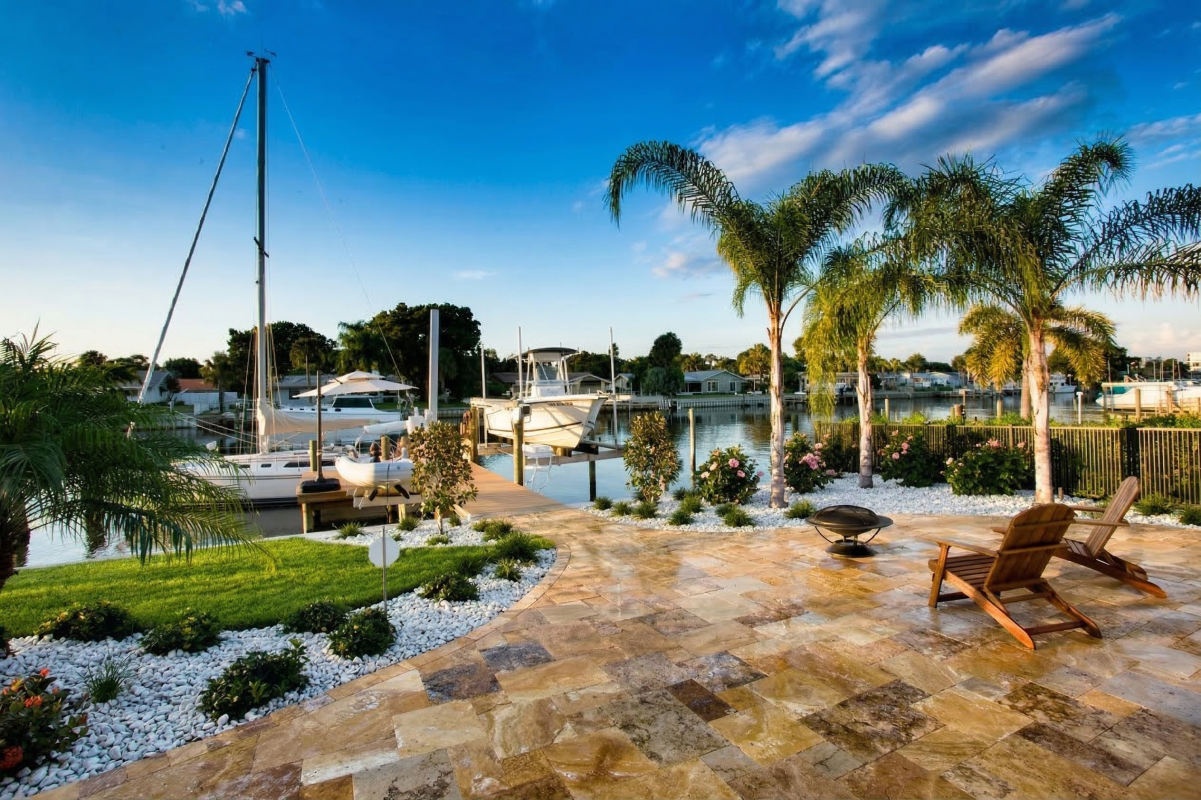 A luxurious waterfront backyard in St. Petersburg, Florida, featuring a custom travertine stone patio, tropical landscaping with palm trees, white river rock beds, a fire pit, and wooden Adirondack chairs overlooking a private boat dock.