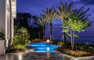 Luxury Clearwater Beach waterfront landscape architecture featuring a custom travertine pool deck, gas fire pit, and professional palm tree uplighting.